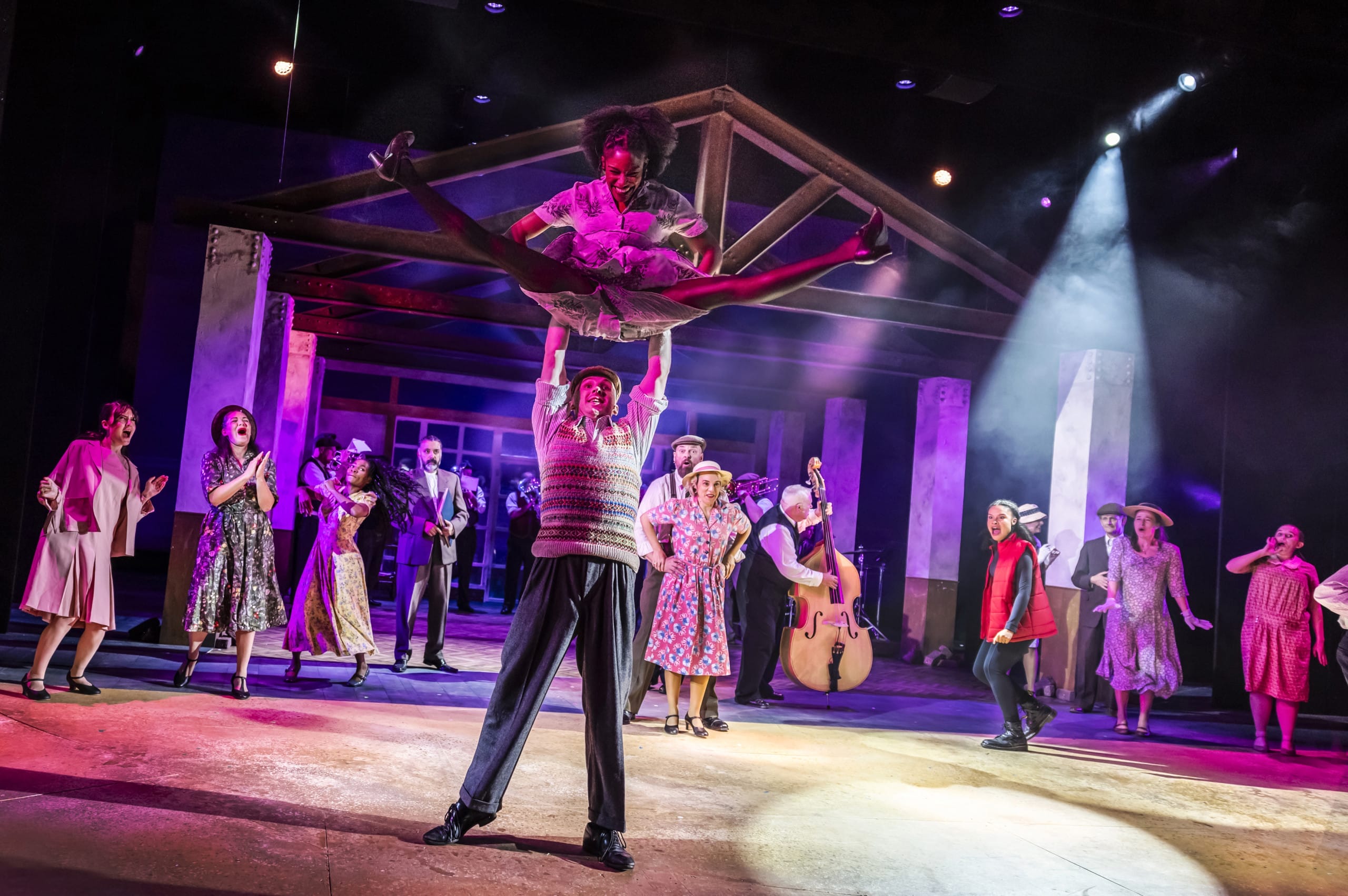 Cast members dancing on the stage in purple lighting. 2 main characters are in the centre, the man holding the woman up in the air while she is mid jump in a splits position