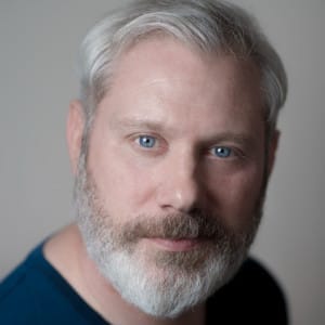 A close-up headshot of a man with light gray hair and a full beard, looking directly into the camera with piercing blue eyes. He has a calm expression, wearing a dark shirt, with a neutral background that highlights his facial features.