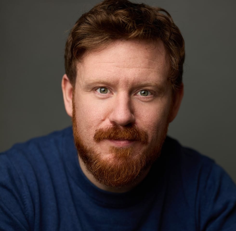 This image is a close-up portrait of John Holt-Roberts with short, tousled hair and a full beard, wearing a blue sweater. The individual has a warm expression with direct eye contact, and the lighting highlights their facial features, particularly the texture of the beard and the clarity of their eyes. The background is a solid, neutral grey, ensuring that the focus remains on the subject's face.