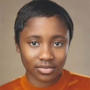 This image is a close-up portrait of Mya Fox Scott with short, smooth hair styled close to the head. The individual is wearing an orange velvet top and has a neutral expression, with direct eye contact and minimal makeup, highlighting their natural features. The background is a soft, neutral tone, which keeps the focus on the subject's face.