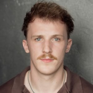 A headshot of a young man with short, wavy brown hair and a light mustache, looking directly into the camera with a neutral expression. He is wearing a brown shirt and a thin silver chain necklace, with a dark, textured background adding contrast to his features.