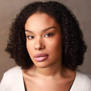 This image is a close-up portrait of Tiana Maria Harrison with medium-length curly hair, wearing a white ribbed cardigan over a black top. The individual has a calm and composed expression, with direct eye contact and natural makeup that highlights their smooth complexion. The background is soft and neutral, ensuring the focus remains on the subject's face.