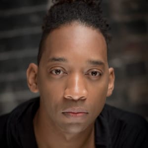 This image is a close-up portrait of Oraine Johnson with dark hair styled in a top knot, wearing a black top. The individual has a serious expression, with direct eye contact, and the lighting accentuates the smooth texture of their skin. The background features a dark, blurred brick wall, adding a subtle contrast that draws attention to the subject's face.