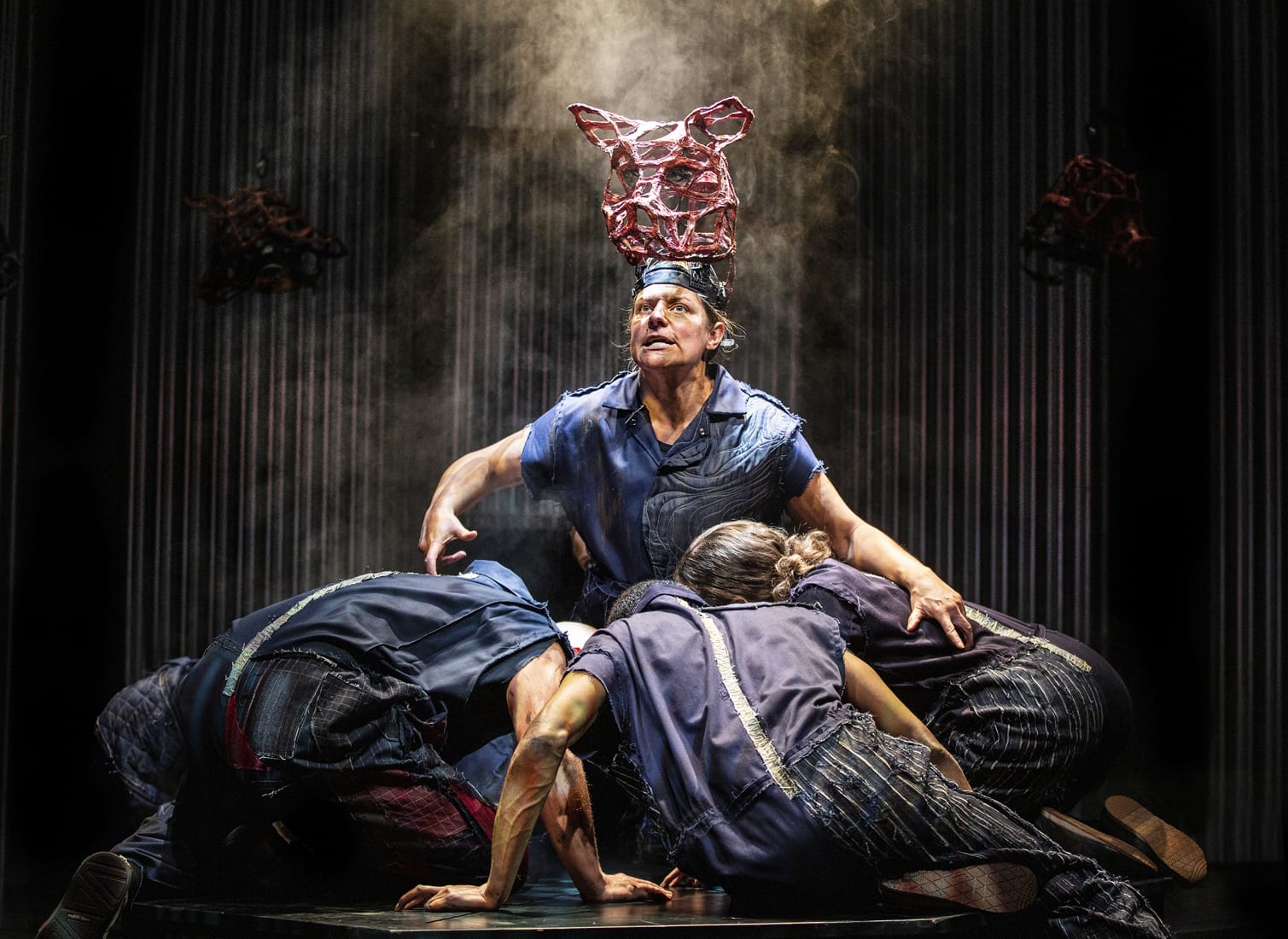 A dramatic stage scene from a production of Animal Farm featuring a central figure wearing a sculptural headpiece resembling a pig, illuminated by theatrical lighting. Surrounding the figure, other performers huddle on the floor in distressed, patchwork-style costumes, creating a tense and atmospheric moment.