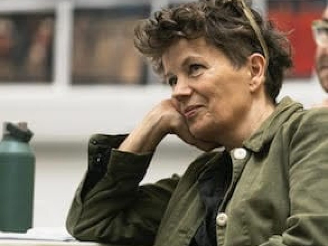 A candid photo of Sally Cookson, the theatre director of "Wonder Boy," sitting at a table with her head resting on her hand. She is wearing a green jacket and has short, curly hair. In the background, there are blurred images on the wall and a water bottle on the table.