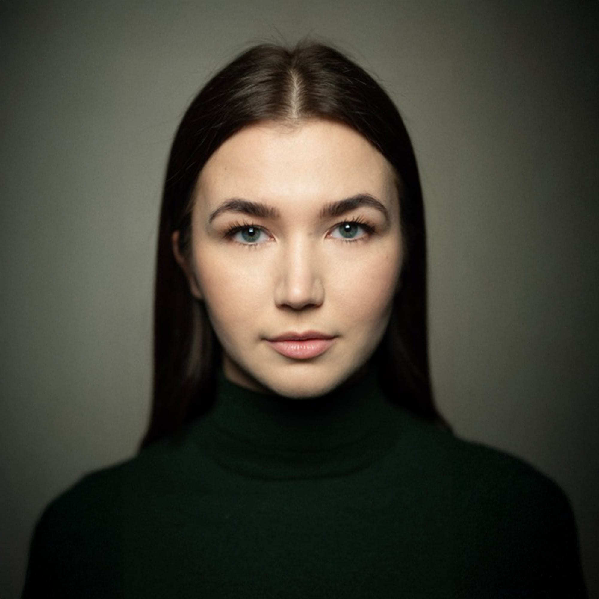 Studio portrait of a young woman with long, straight brown hair, wearing a dark green turtleneck. She looks confidently into the camera with a calm, poised expression against a softly lit, neutral background.