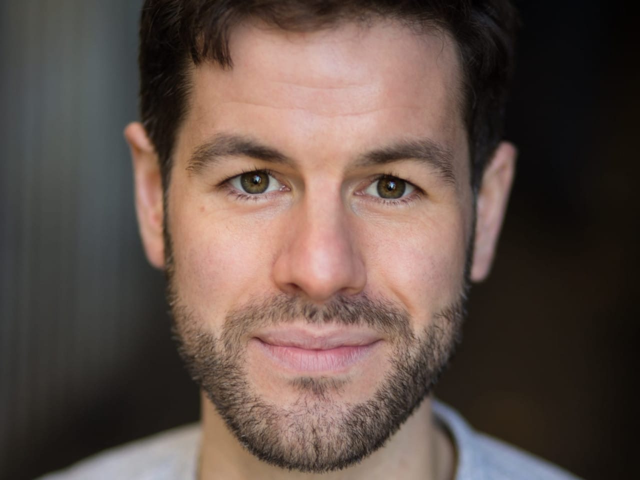 Performer’s headshot - low cut dark brown hair with a beard and light blue striped shirt on