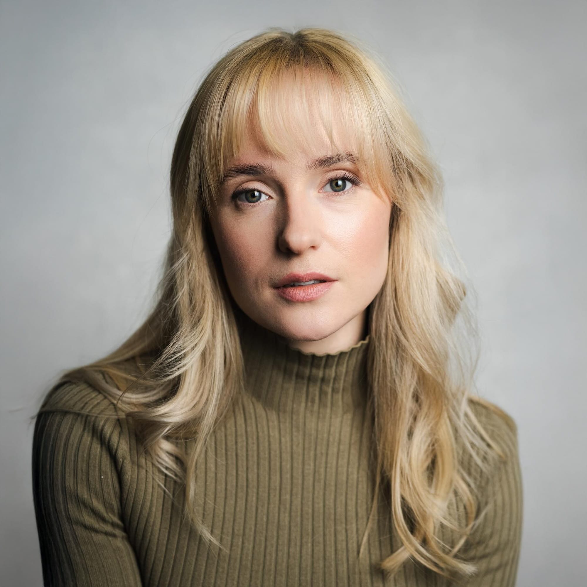 Portrait of a woman with long, wavy blonde hair and light bangs, wearing a ribbed olive-green turtleneck top. She gazes softly at the camera with a calm and thoughtful expression, set against a light grey background.