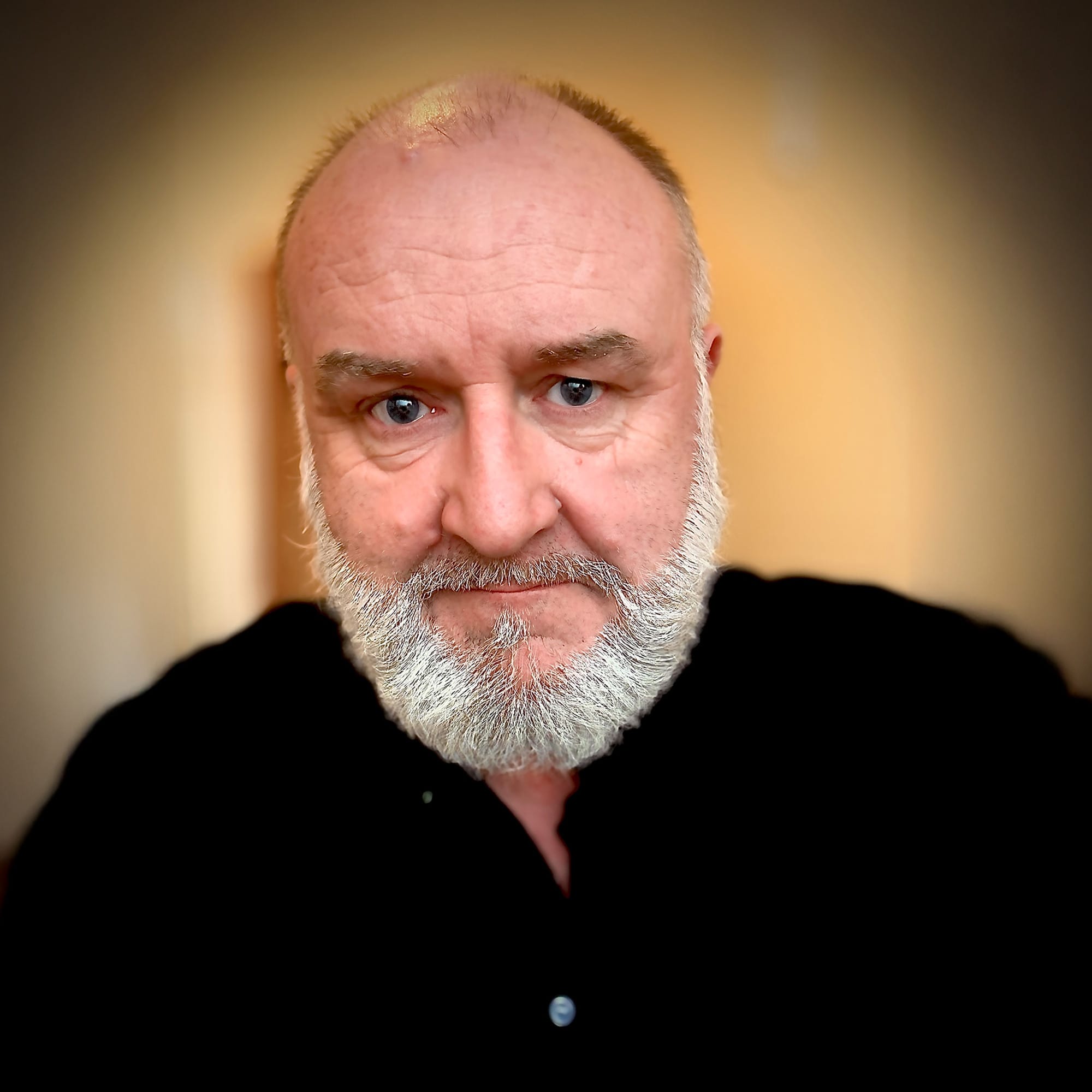 Close-up portrait of an older man with a bald head, full white beard, and piercing blue eyes, wearing a black button-up shirt. The background is softly blurred in warm tones, drawing focus to his serious, direct expression.