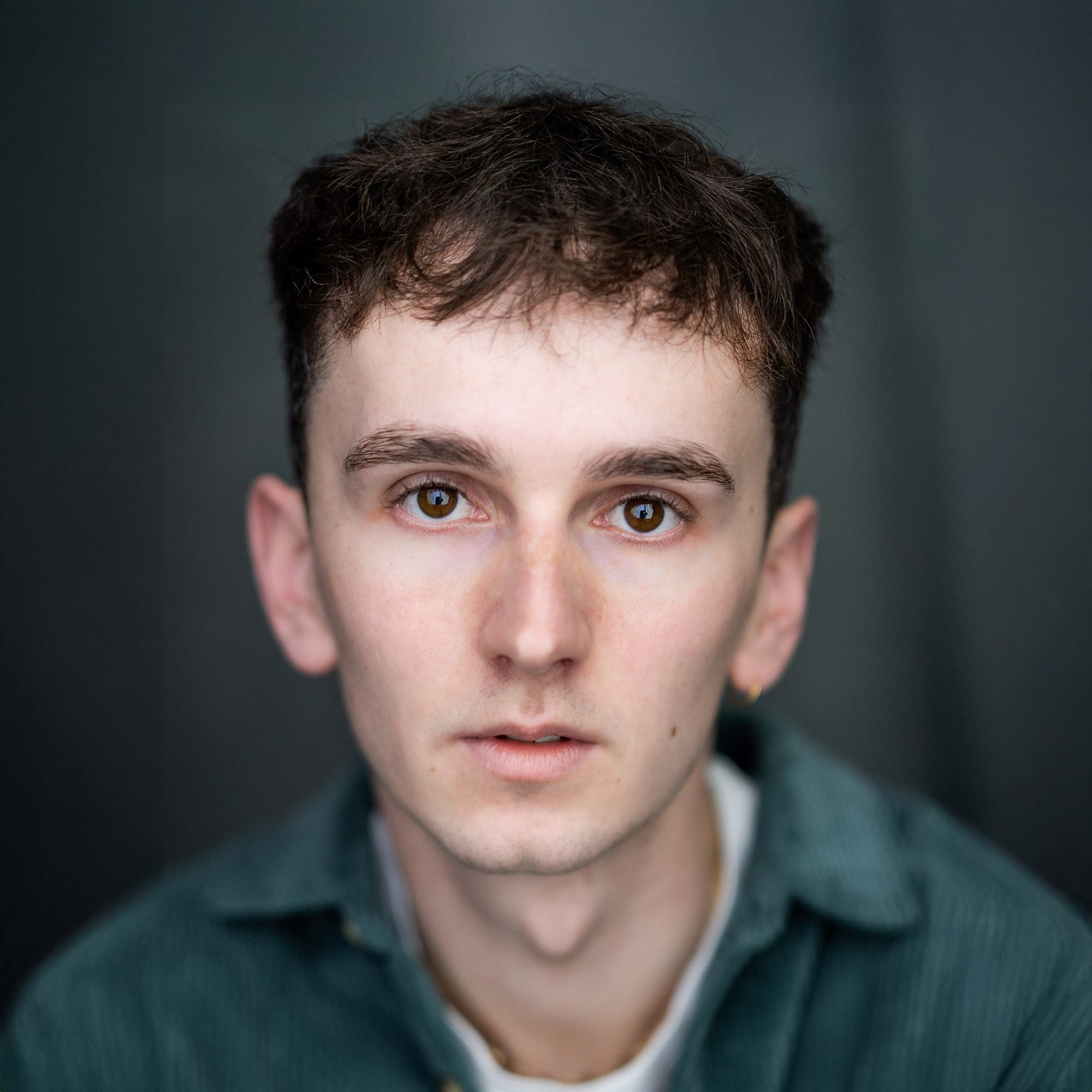 Portrait of a young man with short, tousled dark brown hair and warm hazel eyes, wearing a green striped shirt over a light grey top. He faces the camera with a neutral, slightly intense expression, set against a dark, softly blurred background.