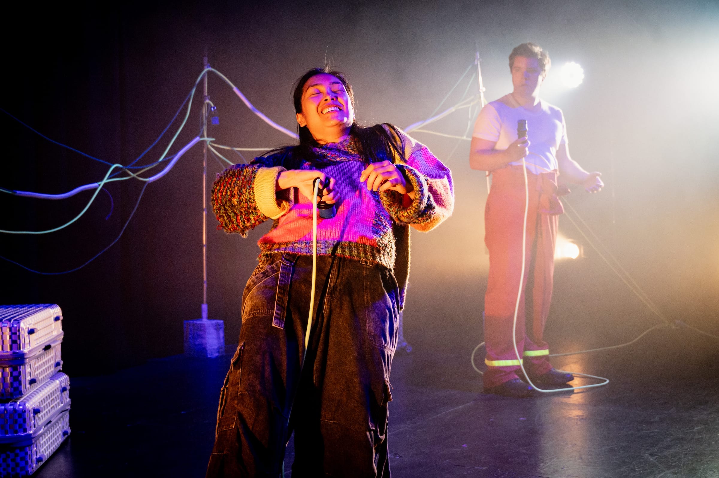 A dynamic production image from Firewall, featuring a performer in a colourful knit sweater, energetically handling a microphone or cable, illuminated by warm, dramatic stage lighting. In the background, another performer in a white shirt and red trousers holds a microphone, surrounded by glowing lights and wires stretching across the stage. The scene exudes energy and creativity, highlighting the innovative set design and engaging performance.