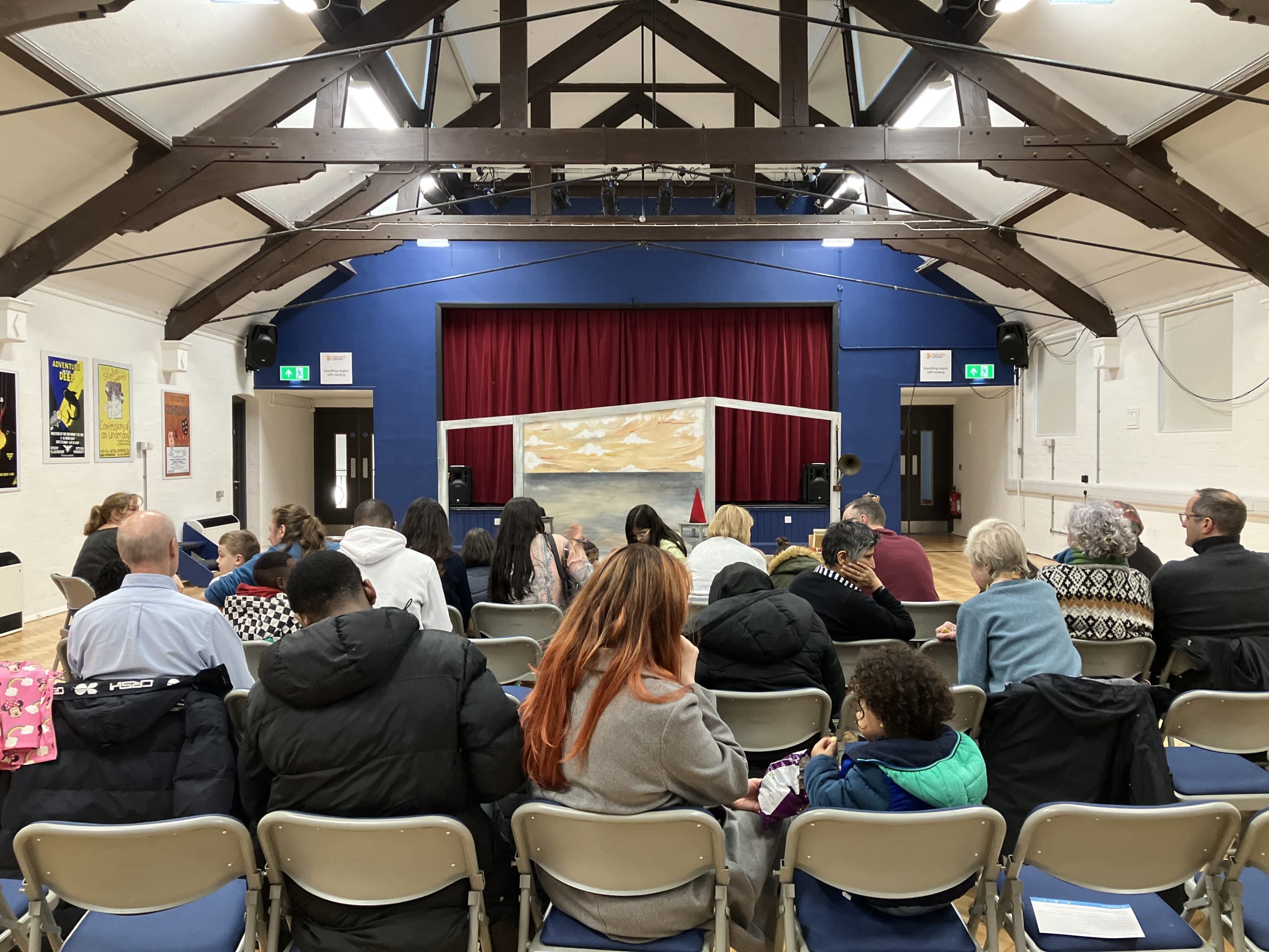An establishing shot of an audience seated in a community hall, awaiting a performance preview of Farmer and the Clown. The stage at the front is framed by a red curtain and features a simple set design with painted scenery depicting a rural landscape. The hall's exposed wooden beams and cosy atmosphere set the tone for an intimate and engaging theatrical event.