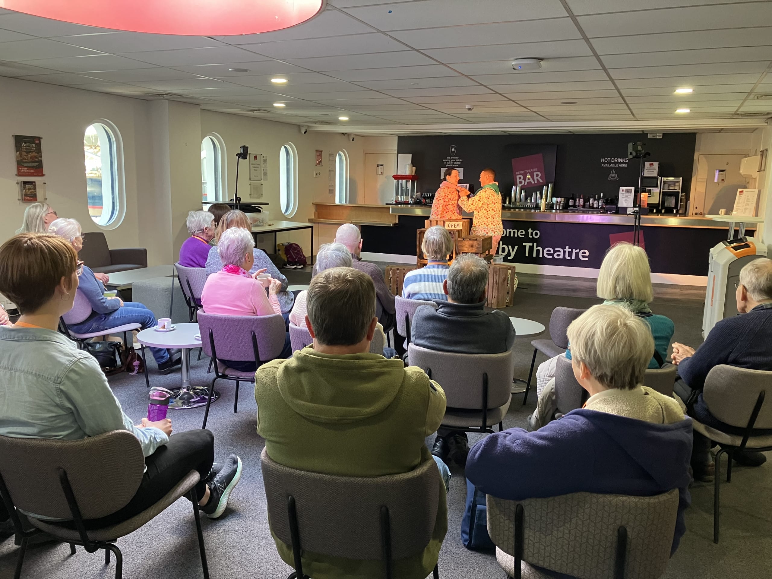 A relaxed setting at the Dementia Café, where an audience of older adults watches a performance of The Gingerbread Man. The performers, dressed in brightly patterned costumes, are animatedly interacting near a cosy bar area with a sign reading 