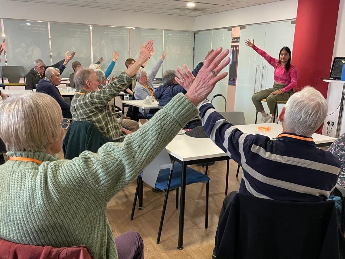 Participants at the Dementia Carers’ Café engage in a movement wellbeing session led by local Movement Director, Aishwarya Deshpande, supported by DT Communities. The group, seated at tables, follows the instructor by raising their arms, creating a sense of connection and activity in a welcoming, brightly lit community space. The atmosphere reflects focus and inclusivity.