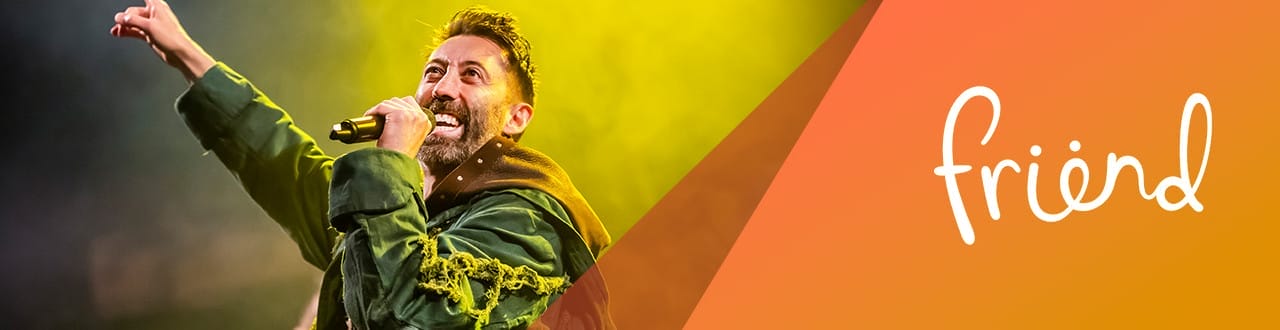 Close-up of a smiling male performer in a green costume passionately singing or speaking on stage with bright yellow lighting. Overlay text on the right reads “friend” in white on an orange gradient background.